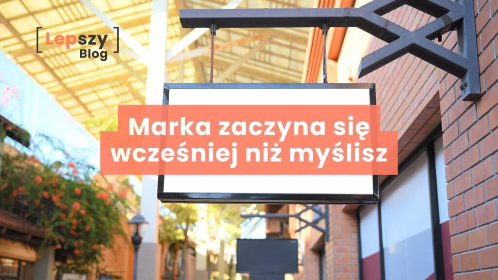 Szyld sklepu zwisający nad ulicą, widoczny na tle miejskiej zabudowy – napis „Marka zaczyna się wcześniej niż myślisz”, symbol pierwszego wrażenia i tożsamości, która buduje się zanim pojawi się produkt czy oferta.